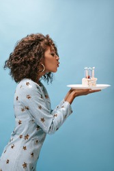 Trendy brunette lady in round modern earrings and blue printed clothes blowing out candles on piece of cake on isolated backdrop..