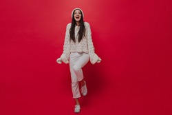 Charming young lady with brunette long hairstyle, bright lips, comfortable knitted clothes, accessories and white jeans, sneakers, smiling and lovely posing against plain red background 