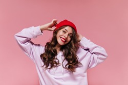 Charming cool girl in purple hoodie puts on red hat. Pretty brunette curly woman smiles widely on pink isolated background.