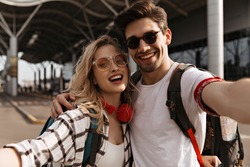 Young attractive blonde woman in plaid shirt and stylish brunette man in sunglasses smiles and takes selfie. Portrait of couple of travelers near airport.