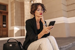 Cool girl with wavy hair in white trousers and balck jacket holding phone outside. Stylish woman in eyeyglases sitting outdoors..