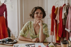 Attractive young curly short-haired woman in floral dress smiles and looks into camera. Portrait of fashion designer in office.