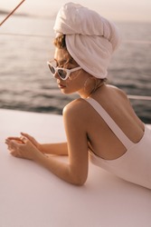 Pretty young girl with light swimsuit, towel on head and stylish sunglasses resting and looking down against background of sunlit sea in summer
