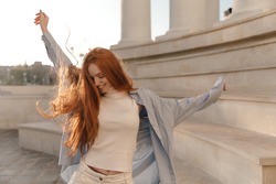 Cute foxy lady dancing outdoors at sunny day. Adorable young long-haired girl , wearing light turtleneck, blue shirt, moving and looking down against city landmark background