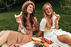 Tanned happy brunette woman in boater and beige polka dot outfit sits on white rug outside. Happy excited blonde lady in sunglasses has picnic with friend and holds piece of melon.