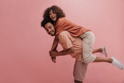 side profile full length cheerful positive cute couple in casual wear. african american man carrying woman smiling with teeth over isolated pastel pink background.