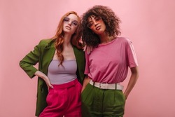 Young fashionable interracial girls in pink and green outfits stand spectacularly against pink wall. Half portrait of red-haired model in jacket, top, pants and brunette in T-shirt and pants with belt