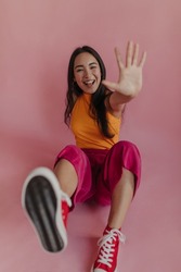 Young beautiful korean girl having fun on pink background while sitting on floor. Happy model with bright appearance in orange T-shirt, crimson pants and red shoes, straightening leg and arm forward.