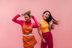 Energetic young two interracial girls dancing side by side and waving hands in studio. Models with light brown and black hair in light spring orange and pink clothes move to music with smile on face.