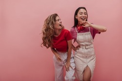 Young caucasian lady laughs hard with her pretty asian girlfriend against pink background. Image of 20s girls pretending dancing to music and having fun with each other in studio.