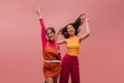 Two beautiful young girls from Ukraine and Asia dancing in pink background with place for text. Blonde and brunette pose with eyes closed, waving hands and ruffling hair, singing and jumping for joy.