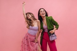 Stylish young interracial white girls listening to music while standing against pink background. Asian model in jacket and raspberry vintage pants stands next to caucasian lady in long skirt and top.