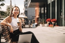 Asian brunette woman with wide smile sitting on bench looks at camera with coffee in her hands on city street. Girl manager in non-office is engaged in additional work at laptop.