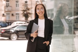 Brunette business young asian woman with laptop in her hands not in full growth smiling at camera. Lady with glasses stands outside office building during her lunch break.