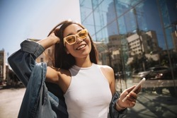 Close-up of fashionably dressed asian tourist in stylish glasses, eyes closed, walking relaxed in center of capital. Brunette in denim jacket with phone in hand. Lifestyle, female beauty concept