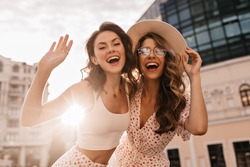 Bottom view of two cheerful beautiful caucasian young women posing for photo in sunbeams. On warm day, brunette and blonde are walking around city in white casual clothes.