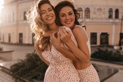 Lovely caucasian young brunette hugs her girlfriend from behind outdoors. Girls smiling with teeth, dressed in white summer clothes. Concept of enjoying weekend, vacation