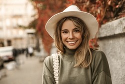Close-up cheerful young european woman smiling with teeth while looking at camera against blurred urban background. Blonde model in hat on autumn day goes to meeting. Lifestyle, female beauty concept