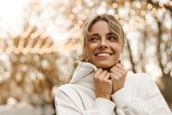 Close-up of pretty young caucasian woman in casual autumn clothes walking in city. Beautiful stylish lady is wrapped white sweatshirt, fashionable hat, autumn fashion trend. Street style concept.