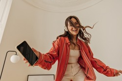 Energetic young model with wireless headphones is having lot of fun in closed white room. Dark-haired girl wear casual clothes holds phone in her hand and smiles cutely at camera.