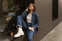 Young caucasian woman using mobile phone listens to music through headphones on street. Sitting on windowsill outside with her eyes closed, girl enjoys moment. Freedom and joy concept