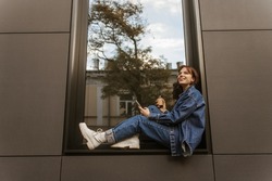 Cheerful pretty young caucasian student sits on windowsill in city, holds phone and coffee. Beauty is dressed denim jacket, jeans and white shoes. Fashion, technology and people concept.