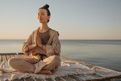 Relaxed young caucasian woman sitting on seashore practices yoga without stress. Model with dark topknot on her head with eyes closed. Cozy beach atmosphere, summer concept.