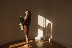 Full length slim caucasian young girl stands sideways holding twisted yoga mat in her hands with space for text. Woman wears sports tight clothes barefoot. Concept of relaxation and meditation.