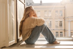 Cute caucasian adult lady with her eyes closed is basking in sun sitting on windowsill on warm day. Long-haired blonde wears casual clothes. Concept of enjoying moment