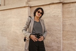 Fashionable adult caucasian brunette woman posing for camera holding her hands on hips against wall on street. Model in sunglasses, gray jacket and black pants. Lifestyle, female beauty concept