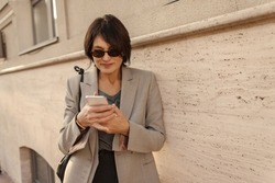 Adult caucasian brunette woman uses modern smart phone while standing outdoors. Model wears sunglasses, grey jacket and shoulder bag. Telephone technology concept