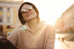 Cheerful adult caucasian brunette lady smiles with teeth while being outdoors on sunny day. Woman wears warm jacket in autumn. Beauty, people emotions and vacation concept.