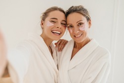 Happy caucasian mother with daughter wearing hydrogel patches smiling looking at camera on white background. Blondes wear robes to take care of their skin. Cosmetic natural trend.