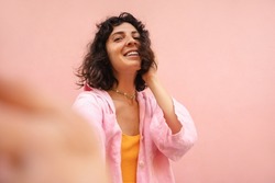 Pretty young brunette caucasian woman takes selfies looking at camera on pink background. Model expresses happiness on casual day, wearing shirt. Summer vacation concept. 
