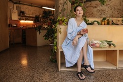 Stylish young caucasian woman looking at camera, holding glass with lemonade sitting indoors. Brunette wears summer blue sundress and black sandals. Lifestyle, different emotions, leisure concept
