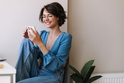 Charming woman holding phone at home . Caucasian smiling brunette woman looking and chatting on smartphone sitting on the floor. Concept of lifestyle, use technology 