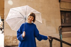 Pretty young caucasian girl with umbrella enjoys spending time outdoors in rainy weather. Short-haired brown-haired woman wears blue autumn sweater. City life concept