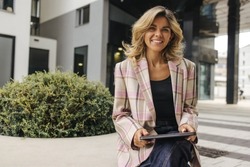 Beautiful young caucasian business woman working on tablet near office. Blonde smiles looking at camera, wears jacket. Concept of use