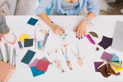 This picture describes the processes of designing clothes. There are hands of a girl drawing sketches on the table. There is creative mess with different stuff on the table.