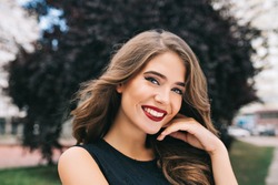 Closeup portrait of pretty girl with long curly hair on street on black trees background. She wears black dress, red lips. She is touching face and smiling to camera.