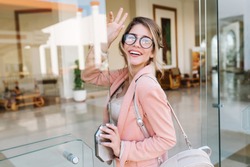 Cheerful pretty young lady entering glass door into modern office, hotel, cafe, business centre and looking back to wave bye. Girl wearing stylish glasses, pink jacket, silver backpack.