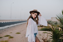 Cute stylish girl in white tunic poses holding her hat next to the road with gray sky on background. Charming young woman in denim shorts and vintage blouse spending time outside enjoys nature views