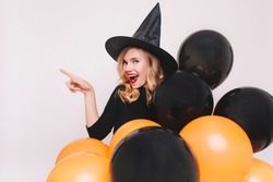 Portrait of blonde woman with surprised smile preparing for halloween party. Indoor photo of curly girl wears black dress and witch hat enjoying themed event.