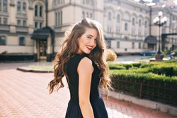 Portrait from back of elegant girl with long curly hair walking on steer on old building background. She has black dress and red lips. She is smiling to camera.