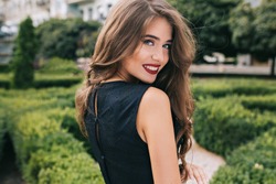 Closeup portrait from back of pretty girl with attractive makeup and red lips on street on green yard background. She wears black dress, has long curly hair. She is smiling to camera.
