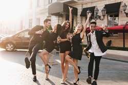 Full-length portrait of fascinating girl in elegant sandals dancing with boyfriend on the street while posing with friends. Outdoor photo of cheerful young people fooling around and drinking champagne