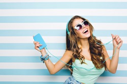 Smiling brown-haired girl enjoying favorite song and dancing in turquoise tank-top. Close-up indoor portrait of excited curly young woman having fun in headphones with phone on striped background.