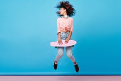 Indoor portrait of african lady with curly hairstyle jumping on blue background during photoshoot. Inspired female model in jeans and black sneakers posing with skateboard.