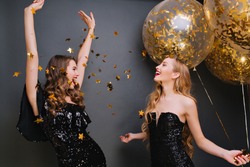 Inspired curly pale woman singing with hands up on dark background. Romantic blonde girl in black outfit holding party balloons and looking at friend which dancing under confetti.