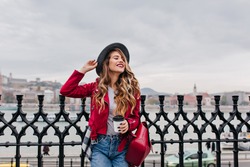 Good-looking girl in jeans and elegant hat posing under gray sky on embankment. Outdoor photo of cute young woman with black manicure drinks coffee on bridge in european city.